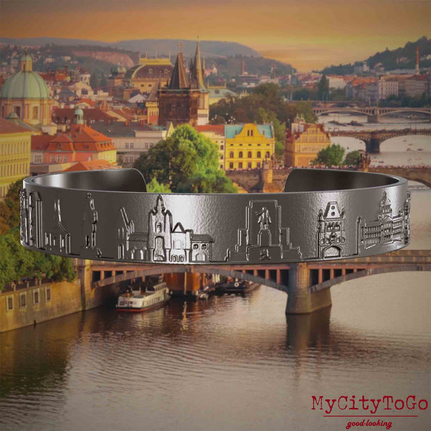 Armreif aus Titan mit Reliefs bekannter Sehenswürdigkeiten der Stadt Prag. Den Hintergrund bildet eine Ansicht der Stadt Prag.