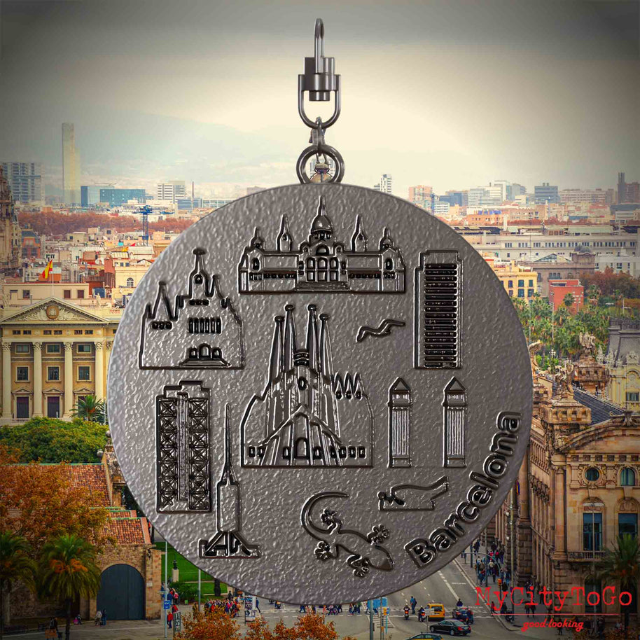 Halskette mit Drehwirbel und Rückseite eines Schmuckanhängers aus Titan, auf dem Reliefs bekannter Sehenswürdigkeiten der Stadt Barcelona abgebildet sind. Den Hintergrund bildet eine Ansicht der Stadt Barcelona.
