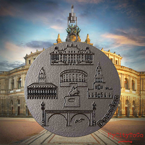 Halskette mit Drehwirbel und Vorderseite eines Schmuckanhängers aus Titan, auf dem Reliefs bekannter Sehenswürdigkeiten der Stadt Dresden abgebildet sind. Den Hintergrund bildet eine Ansicht der Stadt Dresden.