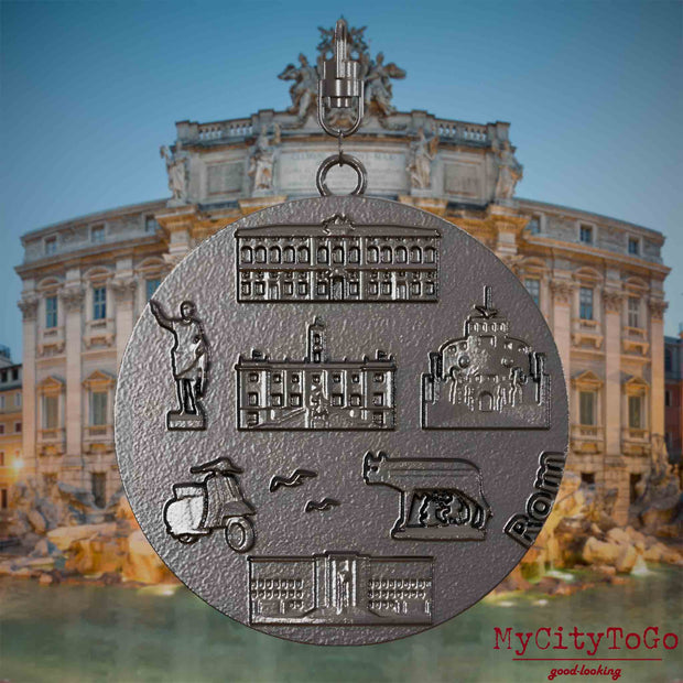 Halskette mit Drehwirbel und Vorderseite eines Schmuckanhängers aus Titan, auf dem Reliefs bekannter Sehenswürdigkeiten der Stadt Rom abgebildet sind. Den Hintergrund bildet eine Ansicht der Stadt Rom.
