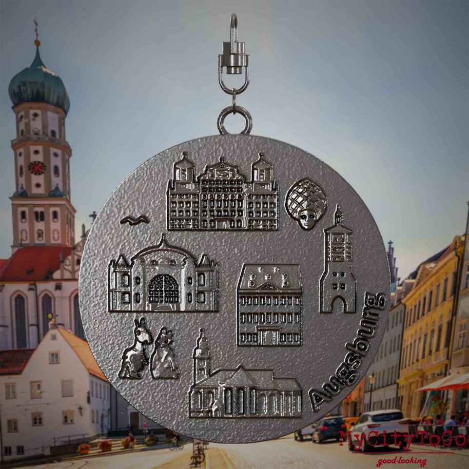 Halskette mit Drehwirbel und Rückseite eines Schmuckanhängers aus Titan, auf dem Reliefs bekannter Sehenswürdigkeiten der Stadt Augsburg abgebildet sind. Den Hintergrund bildet eine Ansicht der Stadt Augsburg .
