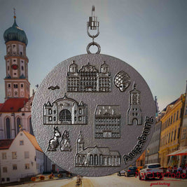 Halskette mit Drehwirbel und Rückseite eines Schmuckanhängers aus Titan, auf dem Reliefs bekannter Sehenswürdigkeiten der Stadt Augsburg abgebildet sind. Den Hintergrund bildet eine Ansicht der Stadt Augsburg .
