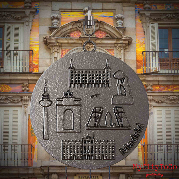 Halskette mit Drehwirbel und Vorderseite eines Schmuckanhängers aus Titan, auf dem Reliefs bekannter Sehenswürdigkeiten der Stadt Madrid abgebildet sind. Den Hintergrund bildet eine Ansicht der Stadt Madrid.
