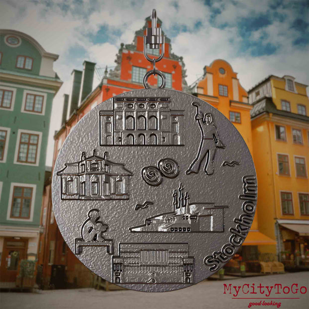 Halskette mit Drehwirbel und Vorderseite eines Schmuckanhängers aus Titan, auf dem Reliefs bekannter Sehenswürdigkeiten der Stadt Stockholm abgebildet sind. Den Hintergrund bildet eine Ansicht der Stadt Stockholm.
