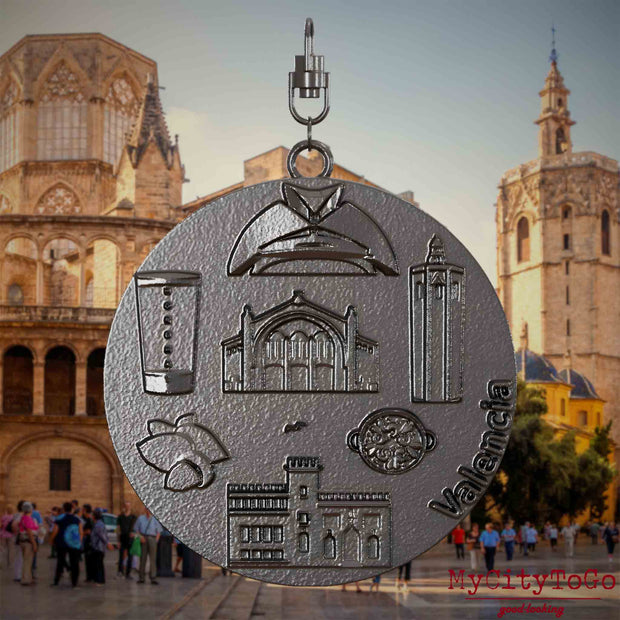 Halskette mit Drehwirbel und Vorderseite eines Schmuckanhängers aus Titan, auf dem Reliefs bekannter Sehenswürdigkeiten der Stadt Valencia abgebildet sind. Den Hintergrund bildet eine Ansicht der Stadt Valencia.
