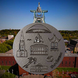 Halskette mit Drehwirbel und Vorderseite eines Schmuckanhängers aus Titan, auf dem Reliefs bekannter Sehenswürdigkeiten der Stadt Bochum abgebildet sind. Den Hintergrund bildet eine Ansicht der Stadt Bochum .
