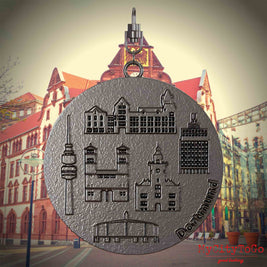 Halskette mit Drehwirbel und Vorderseite eines Schmuckanhängers aus Titan, auf dem Reliefs bekannter Sehenswürdigkeiten der Stadt Dortmund abgebildet sind. Den Hintergrund bildet eine Ansicht der Stadt Dortmund.