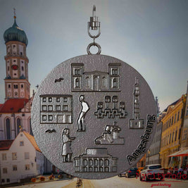 Halskette mit Drehwirbel und Vorderseite eines Schmuckanhängers aus Titan, auf dem Reliefs bekannter Sehenswürdigkeiten der Stadt Augsburg abgebildet sind. Den Hintergrund bildet eine Ansicht der Stadt Augsburg .
