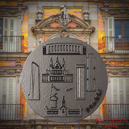 Halskette mit Drehwirbel und Rückseite eines Schmuckanhängers aus Titan, auf dem Reliefs bekannter Sehenswürdigkeiten der Stadt Madrid abgebildet sind. Den Hintergrund bildet eine Ansicht der Stadt Madrid.

