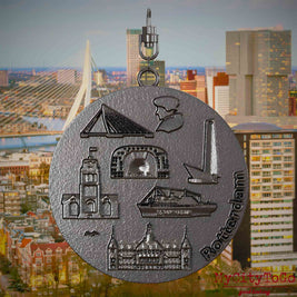 Halskette mit Drehwirbel und Rückseite eines Schmuckanhängers aus Titan, auf dem Reliefs bekannter Sehenswürdigkeiten der Stadt Rotterdam abgebildet sind. Den Hintergrund bildet eine Ansicht der Stadt Rotterdam.
