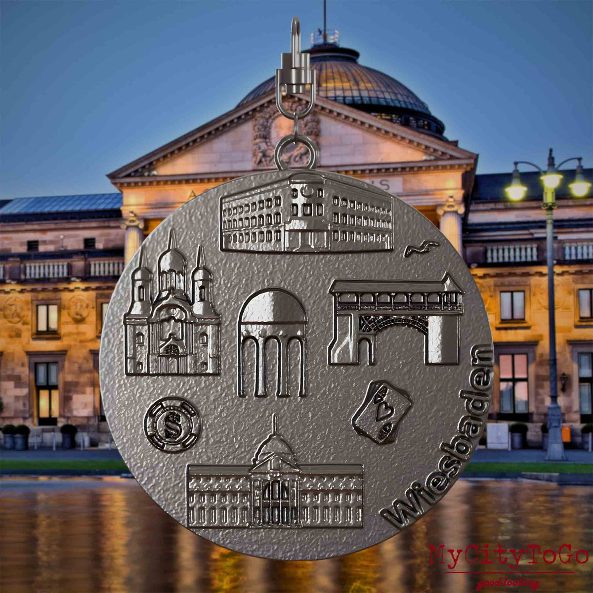 Halskette mit Drehwirbel und Vorderseite eines Schmuckanhängers aus Titan, auf dem Reliefs bekannter Sehenswürdigkeiten der Stadt Wiesbaden abgebildet sind. Den Hintergrund bildet eine Ansicht der Stadt Wiesbaden.
