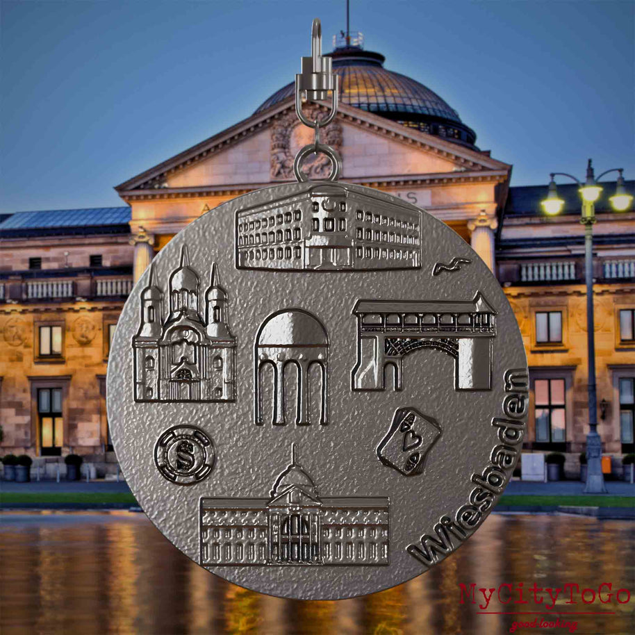 Halskette mit Drehwirbel und Vorderseite eines Schmuckanhängers aus Titan, auf dem Reliefs bekannter Sehenswürdigkeiten der Stadt Wiesbaden abgebildet sind. Den Hintergrund bildet eine Ansicht der Stadt Wiesbaden.
