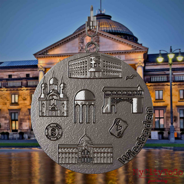 Halskette mit Drehwirbel und Vorderseite eines Schmuckanhängers aus Titan, auf dem Reliefs bekannter Sehenswürdigkeiten der Stadt Wiesbaden abgebildet sind. Den Hintergrund bildet eine Ansicht der Stadt Wiesbaden.
