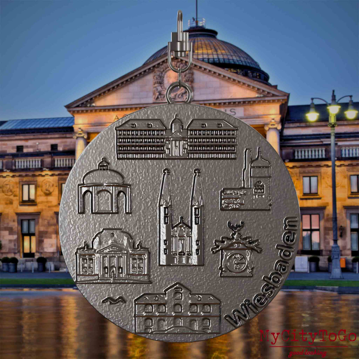 Halskette mit Drehwirbel und Rückseite eines Schmuckanhängers aus Titan, auf dem Reliefs bekannter Sehenswürdigkeiten der Stadt Wiesbaden abgebildet sind. Den Hintergrund bildet eine Ansicht der Stadt Wiesbaden.
