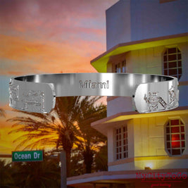 Innenseite eines silbernen Armreifs mit Reliefs bekannter Sehenswürdigkeiten der Stadt Miami und einer Gravur des Stadtnamens. Den Hintergrund bildet eine Ansicht der Stadt Miami.