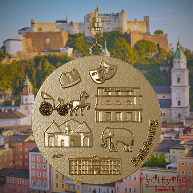 Halskette mit Drehwirbel und Rückseite eines Schmuckanhängers aus vergoldetem Edelstahl, auf dem Reliefs bekannter Sehenswürdigkeiten der Stadt Salzburg abgebildet sind. Den Hintergrund bildet eine Ansicht der Stadt Salzburg.
