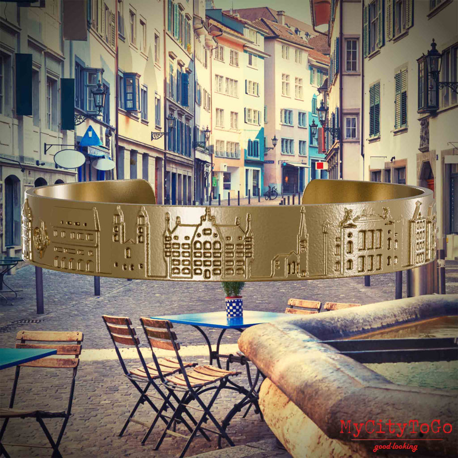Armreif aus vergoldetem Edelstahl mit Reliefs bekannter Sehenswürdigkeiten der Stadt Zuerich. Den Hintergrund bildet eine Ansicht der Stadt Zuerich.