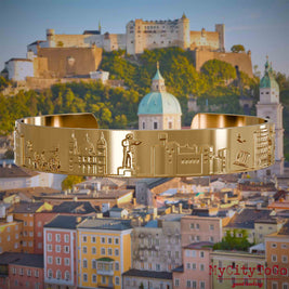 Vergoldeter Armreif mit Reliefs bekannter Sehenswürdigkeiten der Stadt Salzburg. Den Hintergrund bildet eine Ansicht der Stadt Salzburg.