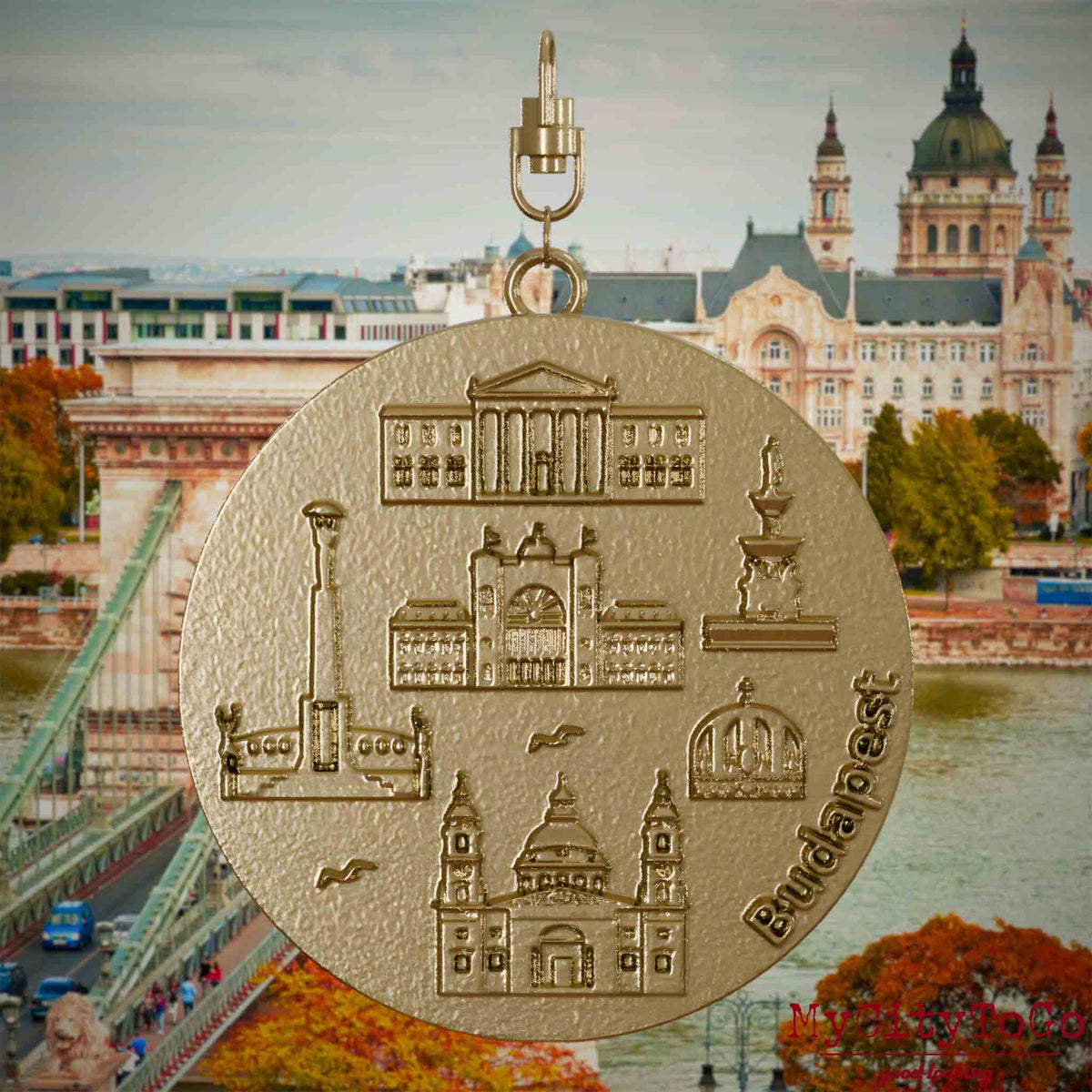 Halskette mit Drehwirbel und Rückseite eines Schmuckanhängers aus vergoldetem Edelstahl, auf dem Reliefs bekannter Sehenswürdigkeiten der Stadt Budapest abgebildet sind. Den Hintergrund bildet eine Ansicht der Stadt Budapest.
