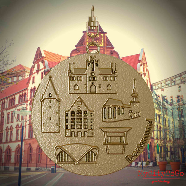 Halskette mit Drehwirbel und Rückseite eines Schmuckanhängers aus vergoldetem Edelstahl, auf dem Reliefs bekannter Sehenswürdigkeiten der Stadt Dortmund abgebildet sind. Den Hintergrund bildet eine Ansicht der Stadt Dortmund.