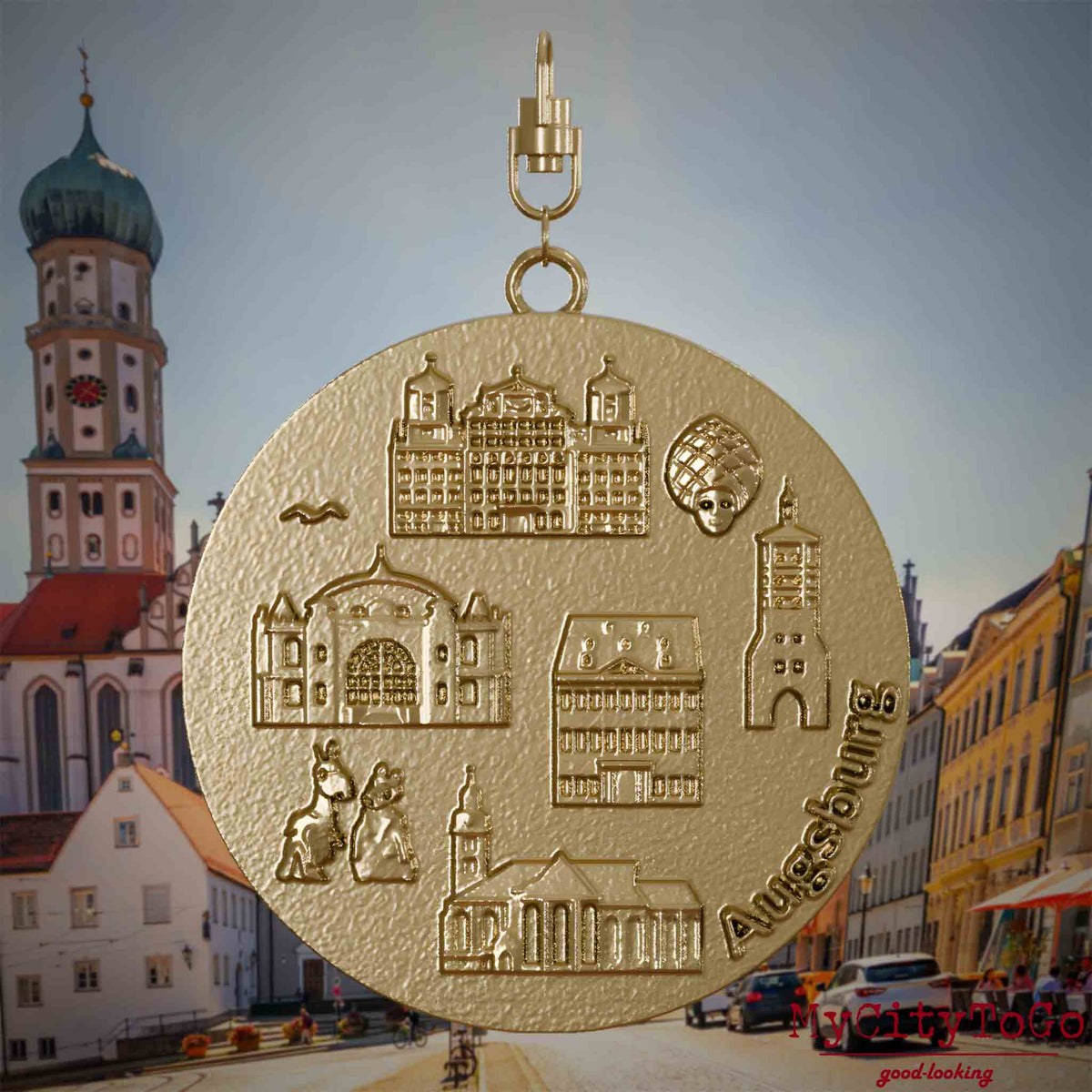 Halskette mit Drehwirbel und Rückseite eines Schmuckanhängers aus vergoldetem Edelstahl, auf dem Reliefs bekannter Sehenswürdigkeiten der Stadt Augsburg abgebildet sind. Den Hintergrund bildet eine Ansicht der Stadt Augsburg.
