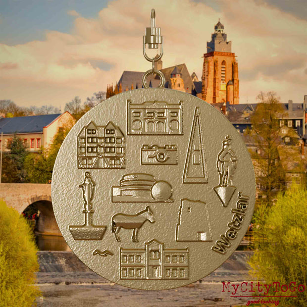 Halskette mit Drehwirbel und Vorderseite eines Schmuckanhängers aus vergoldetem Edelstahl, auf dem Reliefs bekannter Sehenswürdigkeiten der Stadt Wetzlar abgebildet sind. Den Hintergrund bildet eine Ansicht der Stadt Wetzlar.

