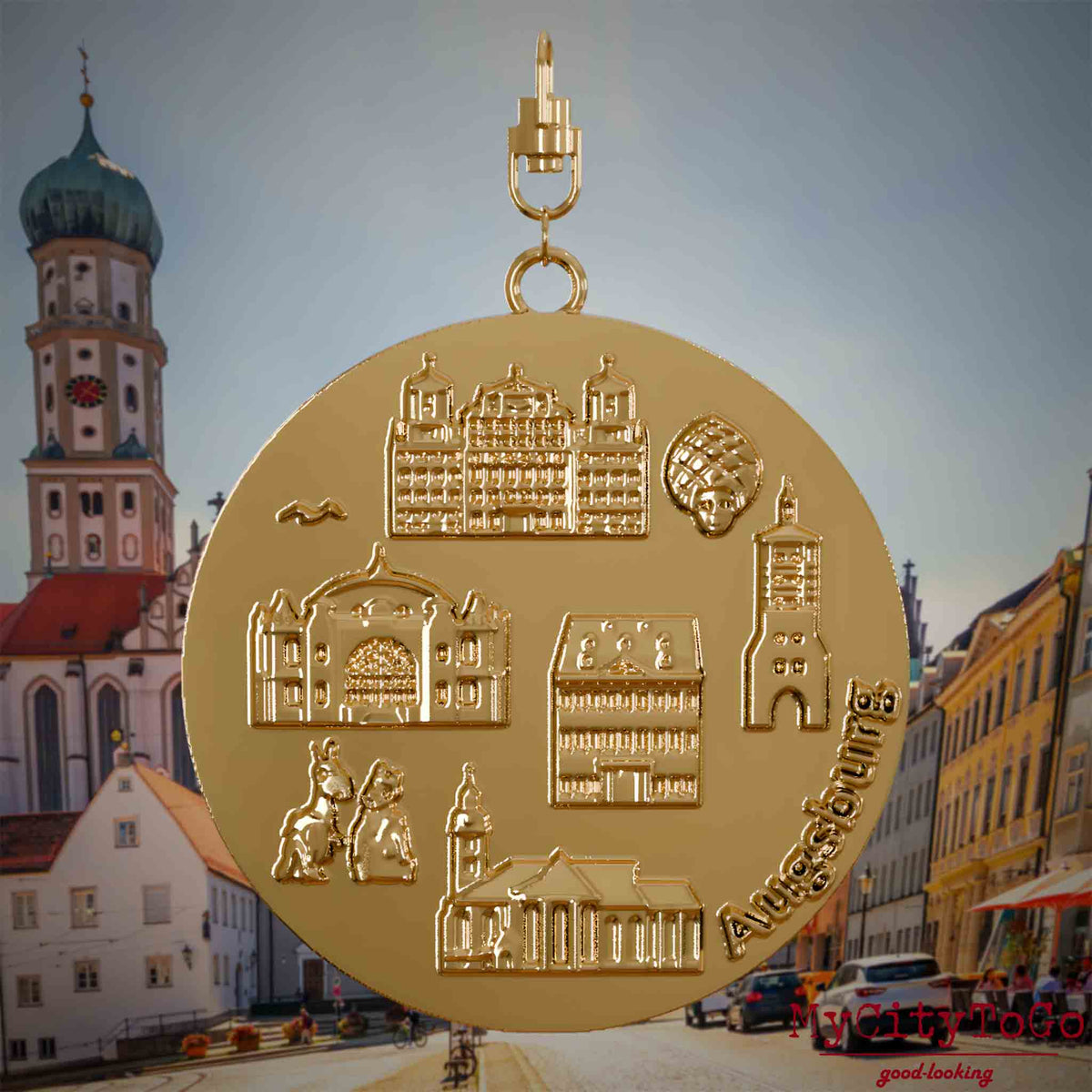 Halskette mit Drehwirbel und Rückseite eines vergoldeten Schmuckanhängers, auf dem Reliefs bekannter Sehenswürdigkeiten der Stadt Augsburg abgebildet sind. Den Hintergrund bildet eine Ansicht der Stadt Augsburg .
