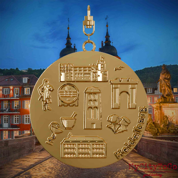 Halskette mit Drehwirbel und Vorderseite eines vergoldeten Schmuckanhängers, auf dem Reliefs bekannter Sehenswürdigkeiten der Stadt Heidelberg abgebildet sind. Den Hintergrund bildet eine Ansicht der Stadt Heidelberg .
