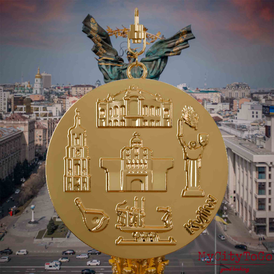 Halskette mit Drehwirbel und Vorderseite eines vergoldeten Schmuckanhängers, auf dem Reliefs bekannter Sehenswürdigkeiten der Stadt Kyjiw abgebildet sind. Den Hintergrund bildet eine Ansicht der Stadt Kyjiw.
