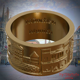Vergoldeter Ring mit Reliefs bekannter Sehenswürdigkeiten der Stadt München und einer Gravur des Stadtnamens auf der Innenseite. Den Hintergrund bildet eine Ansicht der Stadt Muenchen.