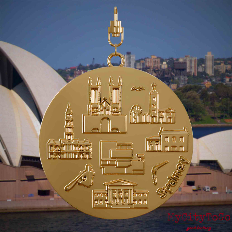Halskette mit Drehwirbel und Rückseite eines vergoldeten Schmuckanhängers, auf dem Reliefs bekannter Sehenswürdigkeiten der Stadt Sydney abgebildet sind. Den Hintergrund bildet eine Ansicht der Stadt Sydney.
