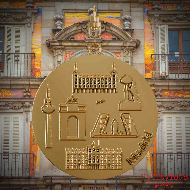 Halskette mit Drehwirbel und Vorderseite eines vergoldeten Schmuckanhängers, auf dem Reliefs bekannter Sehenswürdigkeiten der Stadt Madrid abgebildet sind. Den Hintergrund bildet eine Ansicht der Stadt Madrid.
