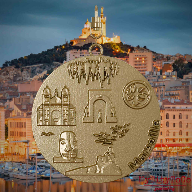 Halskette mit Drehwirbel und Rückseite eines Schmuckanhängers aus vergoldetem Edelstahl, auf dem Reliefs bekannter Sehenswürdigkeiten der Stadt Marseille abgebildet sind. Den Hintergrund bildet eine Ansicht der Stadt Marseille.
