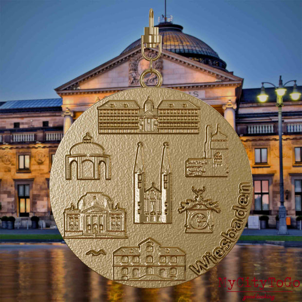 Halskette mit Drehwirbel und Rückseite eines Schmuckanhängers aus vergoldetem Edelstahl, auf dem Reliefs bekannter Sehenswürdigkeiten der Stadt Wiesbaden abgebildet sind. Den Hintergrund bildet eine Ansicht der Stadt Wiesbaden.
