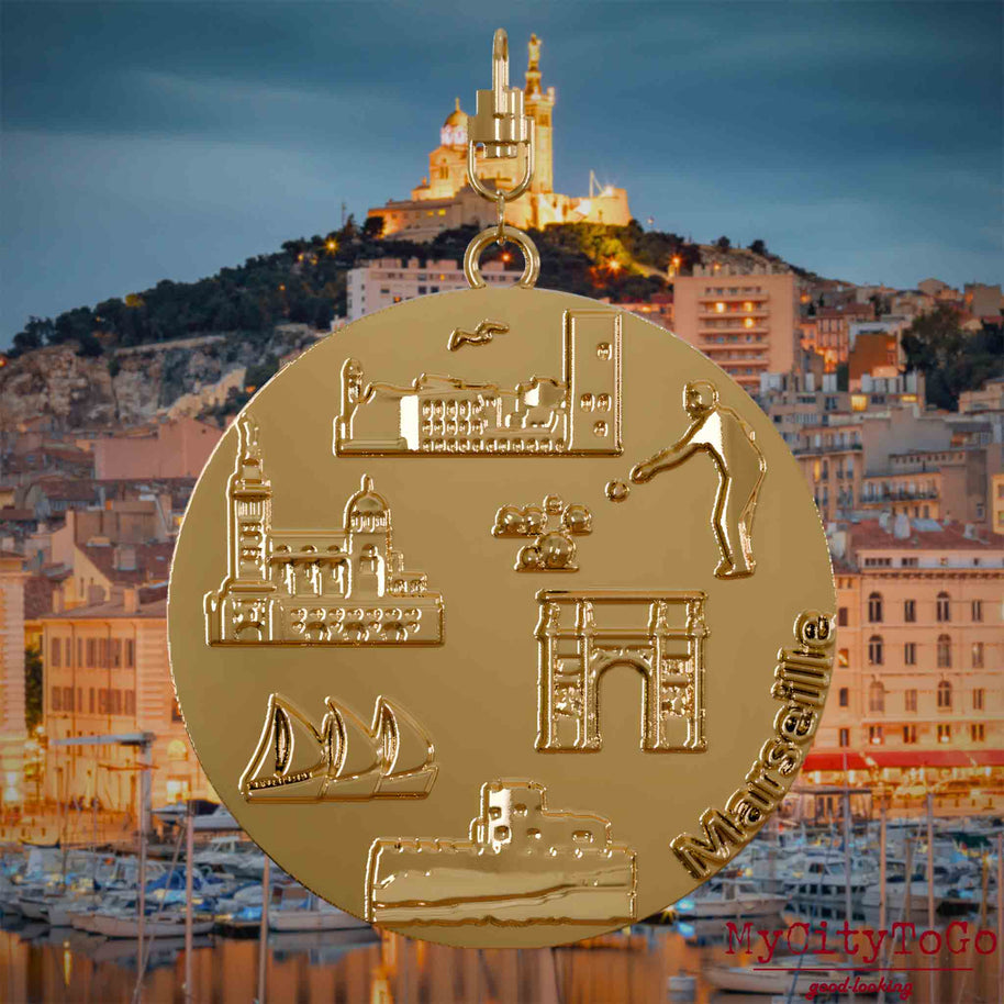 Halskette mit Drehwirbel und Vorderseite eines vergoldeten Schmuckanhängers, auf dem Reliefs bekannter Sehenswürdigkeiten der Stadt Marseille abgebildet sind. Den Hintergrund bildet eine Ansicht der Stadt Marseille.
