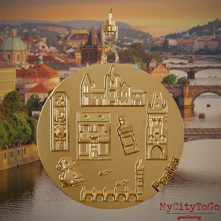 Halskette mit Drehwirbel und Vorderseite eines vergoldeten Schmuckanhängers, auf dem Reliefs bekannter Sehenswürdigkeiten der Stadt Prag abgebildet sind. Den Hintergrund bildet eine Ansicht der Stadt Prag.
