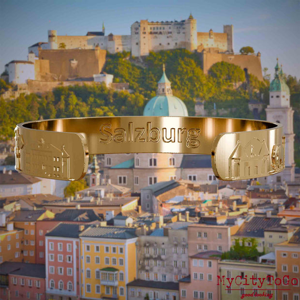 Innenseite eines vergoldeten Armreifs mit Reliefs bekannter Sehenswürdigkeiten der Stadt Salzburg und einer Gravur des Stadtnamens. Den Hintergrund bildet eine Ansicht der Stadt Salzburg.